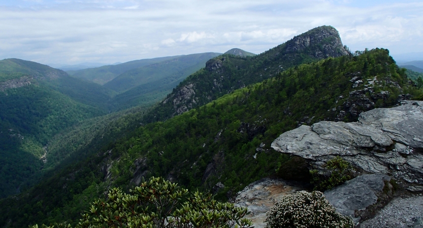 In the foreground, a rocky and green-covered slope jut upwards. Behind the rock formation, vast green mountains appear under a blue and cloudy sky.